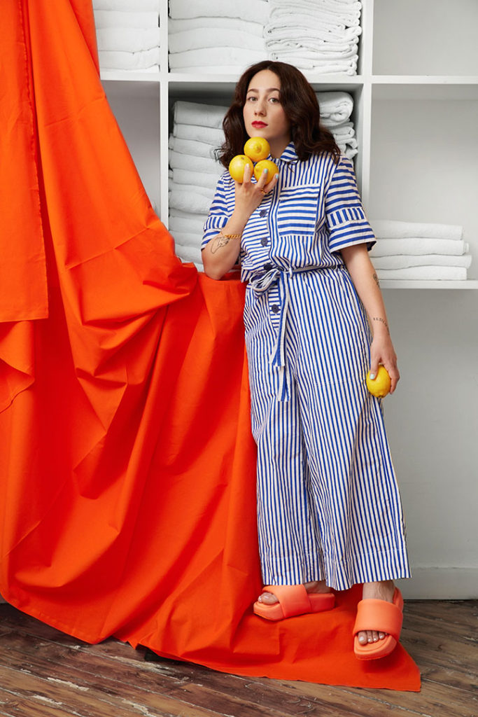 Creative graphic designer Djelissa Latini holding lemons in her hand while standing in front of an orange backdrop while wearing a blue and white striped outfit and orange platform sandals.
