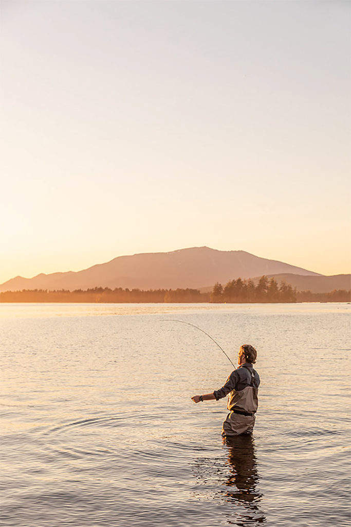 Documentary style photography of a man fishing in water up to his waist during sunrise, with mountains in the distance. By Greta Rybus.