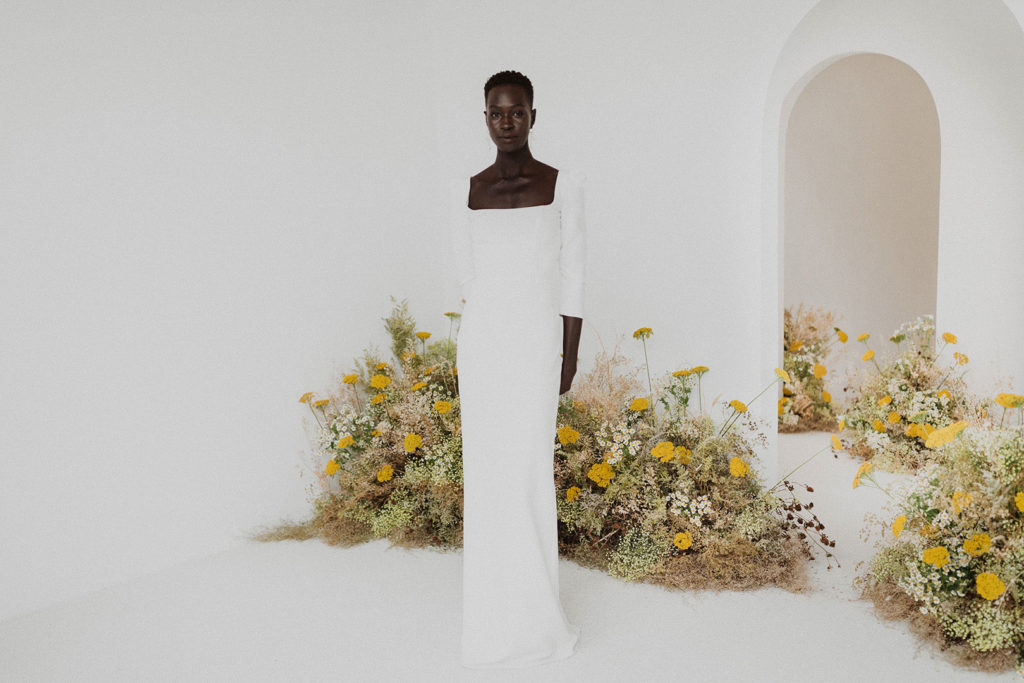 Modern portrait of a slender POC woman in a white maxi dress. Behind her, light green and yellow flowers frame white walls with arched doorways.