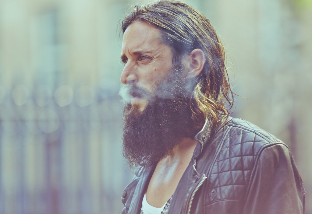 Stylistic portrait of a bearded guy with shoulder length brown hair dressed in a leather jacket and squinting while exhaling smoke. 