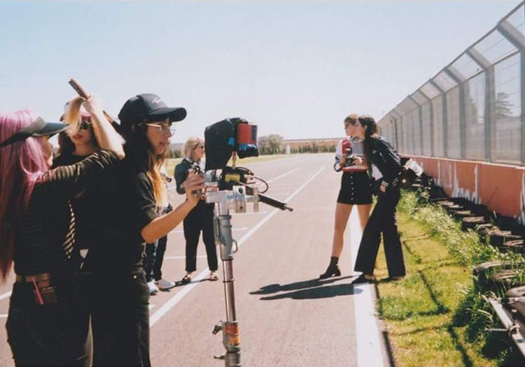 Behind the scenes photograph of award-winning photographer and director, Kell Mitchell. Mitchell is standing on a running track while operating a camera. A hairstylist fixes a model's hair in the background behind the camera.