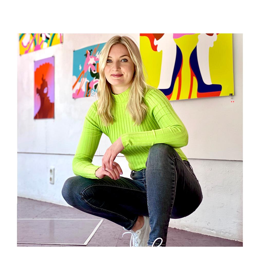 Photo of digital illustrator Lisa Tegtmeier smiling and crouched in front of her creative and colorful illustrations hung on a wall.