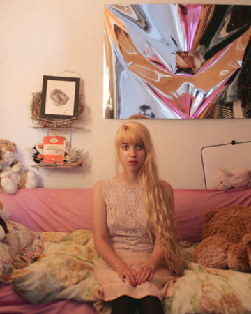 Photo of portrait photographer Lissyelle Laricchia sitting on a couch covered in a pink sheet. Creative and inspiring photographer Lissy Laricchia looking at the camera surrounded by stuffed animals and various art pieces.