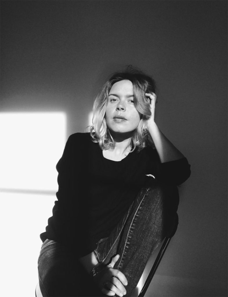 Black and white portrait of creative mixed-media artist, Maria Piessis, leaning casually in a chair while running her hand through her hair.